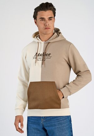 Le sweat à capuche présente un coloris beige et crème en jeu de blocs, une poche avant et le texte imprimé « Atelier » sur la poitrine. Tissu en mélange de coton doux.