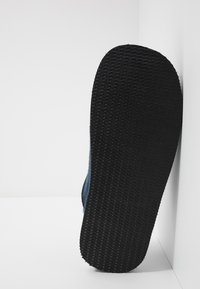 Sort gummisål med et tekstureret grip-mønster, fastgjort til en blå tekstiloverdel, set fra siden mod en ensfarvet baggrund.