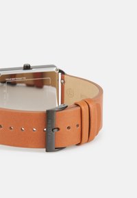 Orologio quadrato in acciaio inossidabile con cinturino in pelle marrone. Il cinturino presenta una finitura testurizzata e una fibbia in metallo con il nome del marchio inciso.