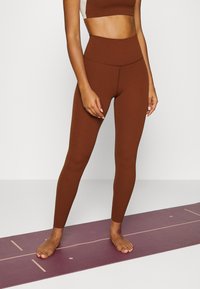 Brown high-waisted leggings made from stretchy fabric, fitted through the legs. Model stands barefoot on a maroon yoga mat with line patterns.