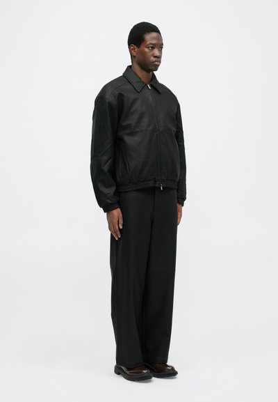 Man die staat in een zwarte zip-up jas met elastische manchetten en zoom, wijde zwarte broek en donkerbruine schoenen tegen een effen witte achtergrond.
