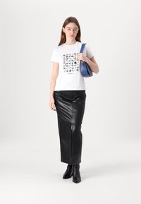 Balta marškinėliai su juodu grafikiniu spaudiniu, derinami su juoda odine sijonu su priekinėmis įpjovomis. Modelis laiko mėlyną rankinę.