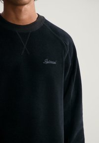 Sudadera de cuello redondo negra llevada por una persona, con pequeño texto en cursiva bordado en el pecho que dice "Dotted".