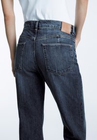 Mörk denimjeans med hög midja, bakfickor och en tan läderlapp på midjebandet. Tyget har en subtil textur och blekning.