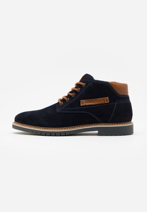 Bottine en daim bleu foncé avec lacets et col marron, coutures claires, semelle en caoutchouc et étiquette "bugatti" sur le côté.