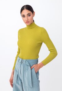 Ribota turtlenecko megztinis ryškiai alyvuogių žalios spalvos, liekna forma su ilgomis rankovėmis, derinamas su aukštu juosmeniu šviesiai mėlynomis kelnėmis, turinčiomis diržą.