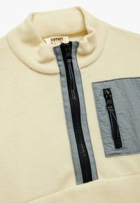 Cremefarbener Fleece-Pullover mit Viertelreißverschluss und grauen Akzenten, ausgestattet mit einer Brusttasche und doppelten Reißverschlüssen. Weiche Textur, hoch geschnittenes Kragen-Design.