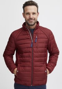 Giacca imbottita rossa con colletto alto, caratterizzata da cuciture orizzontali, chiusura con zip e tirazip blu. Maniche lunghe con polsini elastici.