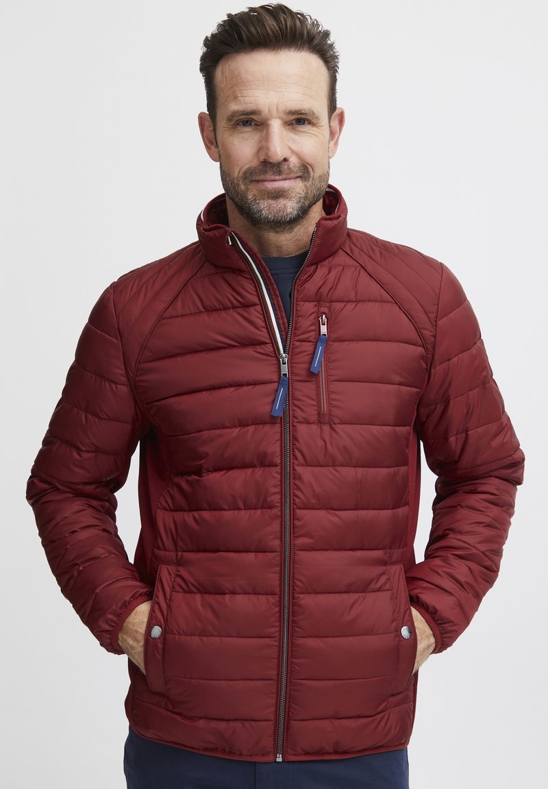 Giacca imbottita rossa con colletto alto, caratterizzata da cuciture orizzontali, chiusura con zip e tirazip blu. Maniche lunghe con polsini elastici.