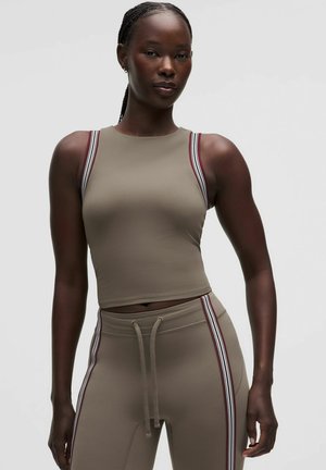 Vrouw staand, gekleed in een aansluitende taupekleurige mouwloze crop top en bijpassende broek met zijstrepen en een taillekoord tegen een effen achtergrond.