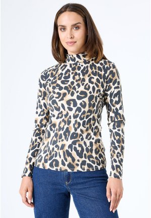 Vrouw in een beige, zwarte en bruine luipaardprint longsleeve top met een hoge hals, gecombineerd met blauwe jeans, staand tegen een witte achtergrond.