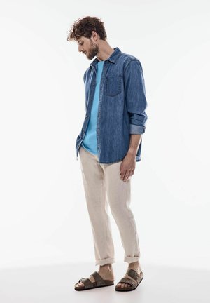 Chemise en jean avec boutons sur un t-shirt bleu clair, associée à un pantalon en lin beige et des sandales marron. Look décontracté, manches retroussées et détails de poche.