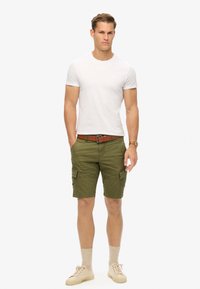 T-shirt bianca a girocollo abbinata a pantaloni cargo verde olivo con tasche laterali, completata da una cintura marrone e sneakers beige.