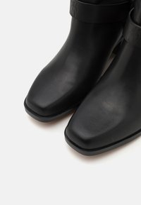 Bottes noires à la cheville en cuir lisse, avec un bout carré et une bande embossée du logo sur le dessus. Semelle plate noire.
