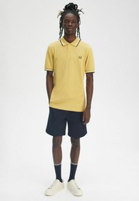 Polo giallo con colletto a contrasto bianco e nero, abbinato a pantaloni corti blu scuro. Presenta sneakers bianche e calze scure con strisce.