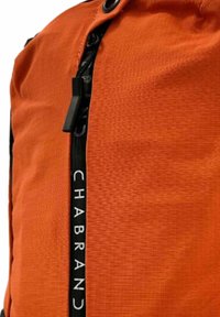 Sac à dos en tissu orange avec une surface texturée, doté d'une fermeture éclair noire centrale et d'une sangle noire marquée sur le côté.