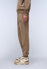 Bruine katoenen joggingbroeken met een ontspannen pasvorm, voorzien van een geribbelde tailleband en een klein logo-opdruk op het been, gecombineerd met bruin en witte sneakers.
