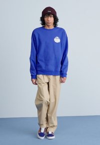 Felpa blu con logo frontale, pantaloni beige larghi e sneakers con motivo. Il modello indossa un berretto di maglia marrone. Sfondo semplice.