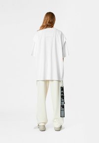 Camiseta blanca de gran tamaño con mangas cortas, parche en la parte posterior, combinada con pantalones de pierna ancha en color crema y zapatos en tonos neutros con suelas texturizadas.