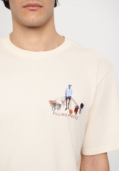 Beige bomull t-skjorte med et grafisk motiv av en person som går med flere hunder, med teksten "FILLING PIECES" under illustrasjonen.