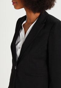 Svart blazer med en skräddarsydd passform, med slagkrage, enkel knappstängning och sidoficka. Bärs över en vit skjorta.