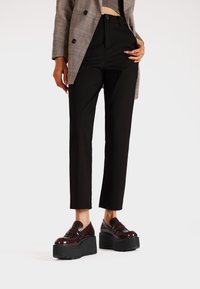 Pantaloni tapered neri con vita alta, abbinati a mocassini in vernice bordeaux con plateau. Giacca con motivo a quadri in tonalità neutre.