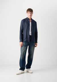 Navy blue linen shirt with a relaxed fit, buttoned down front, and long sleeves, paired with faded denim jeans and white sneakers.