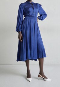Vestido azul plisado con cuello alto, mangas largas y cintura fruncida; combinado con tacones de slingback de punta blanca.