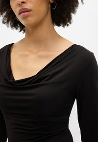 Črn dolg rokav top z drapirano cowl ovratnikom in gladko teksturo. Tkanina se zdi mehka in prilegajoča, kar poudarja silhueto.