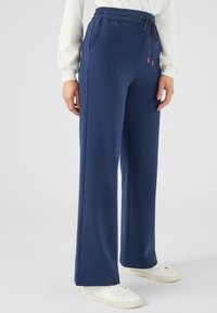 Marineblauwe, wijde broek met een elastische tailleband en trekkoord, met zijzakken en een gestructureerde stof. Draagt met witte sneakers.