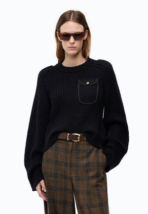 Persona che indossa un maglione nero lavorato a maglia con tasca sul petto, pantaloni marroni a quadri, cintura marrone e occhiali da sole rettangolari marroni, in piedi su sfondo bianco.