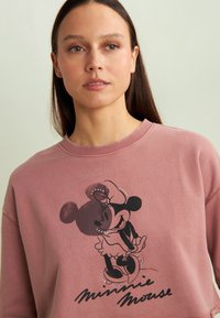 Sudadera rosa con cuello redondo que presenta un gráfico negro de Minnie Mouse y el texto "Minnie Mouse" en una tipografía divertida.