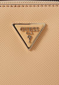 Beige strukturierte Geldbörse mit einem dreieckigen, goldfarbenen Metalllabel, das "GUESS USA" und ein Fragezeichen-Design in der Mitte zeigt.