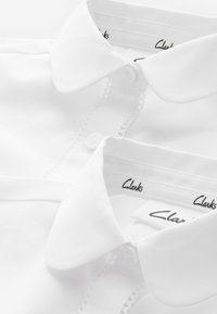 Weißes Hemd mit Kragen, das eine Knopfverschluss und aufwendige Spitzenverzierungen entlang der Knopfleiste aufweist. Markenlabel "Clarks" im Kragen sichtbar.