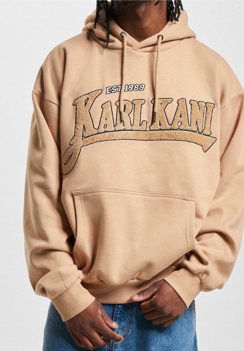 Personne portant un sweat à capuche beige avec le texte « EST 1989 KARL KANI » et un jean bleu, debout devant un fond uni clair.
