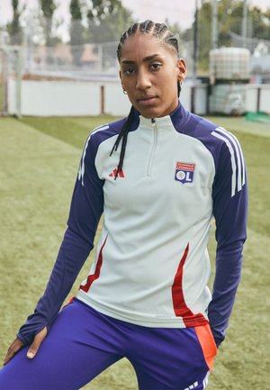 Top atletico bianco e viola con zip a quarto di giro, accenti rossi e logo della squadra. Abbinato a pantaloni viola. Abbigliamento sportivo esposto su un campo d'erba.