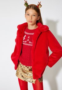 Rød faux fur jakke med krave, rød striktrøje med et juledesign, guldglitrende nederdel og røde strømpebukser. Hårbånd med horn.