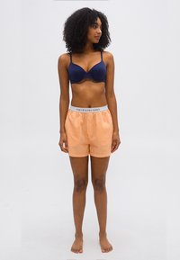 Γυναίκα όρθια, ξυπόλυτη, φοράει μπλε σουτιέν και πορτοκαλί ριγέ μποξεράκια Polo Ralph Lauren, κοιτάζει προς τα αριστερά της σε απλό φόντο.