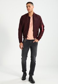 Jachetă bomber bordeaux, material neted, manșete ribbate, buzunar lateral cu fermoar și etichetă roșie; asortată cu o cămașă roz deschis și blugi negri.