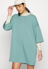 Um vestido de algodão de corte solto e mangas curtas numa tonalidade de teal suave, sobreposto a uma blusa de manga longa com padrão floral, usado com um boné e um colar.