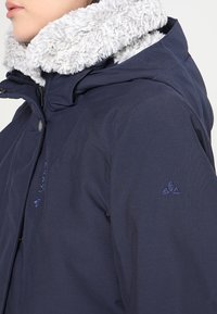 Marinfärgad jacka med en plyschgrå pälskrage, fram zip-stängning och broderad logotyp på ärmen. Tillverkad av ett slätt, hållbart material.