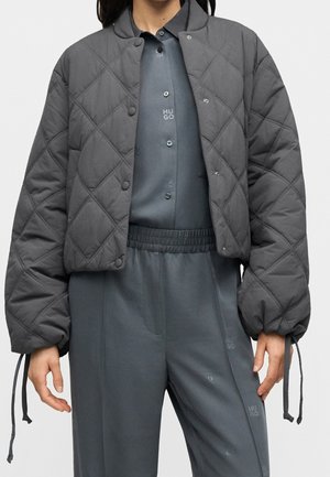 Personne portant une veste matelassée gris foncé par-dessus une chemise boutonnée et un pantalon à taille élastique, debout les mains détendues le long du corps.