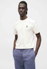 Krémfehér pamut póló kerek nyakkal és rövid ujjakkal. Bal mellrészen kis sárga logóval. Kék melegítőnadrággal párosítva.