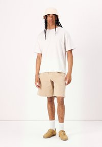 Camiseta blanca de gran tamaño, pantalones cortos beige, gorra de cubo de tono claro, calcetines tobilleros crema y zapatillas color canela, de pie contra un fondo neutral.