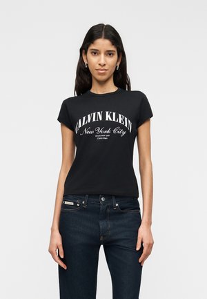 Fiatal nő hosszú, sötét hajjal, fekete Calvin Klein pólóban és sötét farmerben, egy egyszerű, világos háttér előtt állva.