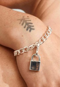 Bracelet en chaîne argentée avec un petit charm en forme de cadenas, avec une finition brillante et un design de maillons texturés, porté sur la peau avec un tatouage visible.