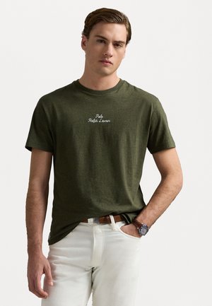 Tricou din bumbac verde măsliniu, cu mâneci scurte, având un logo alb "Polo Ralph Lauren" centrat pe piept. Purtați cu pantaloni albi.