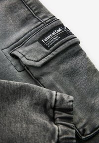 Tejido de mezclilla gris con una textura sutil. Presenta un bolsillo cosido y una etiqueta que dice "Futuro de lo Cool" con instrucciones de cuidado.