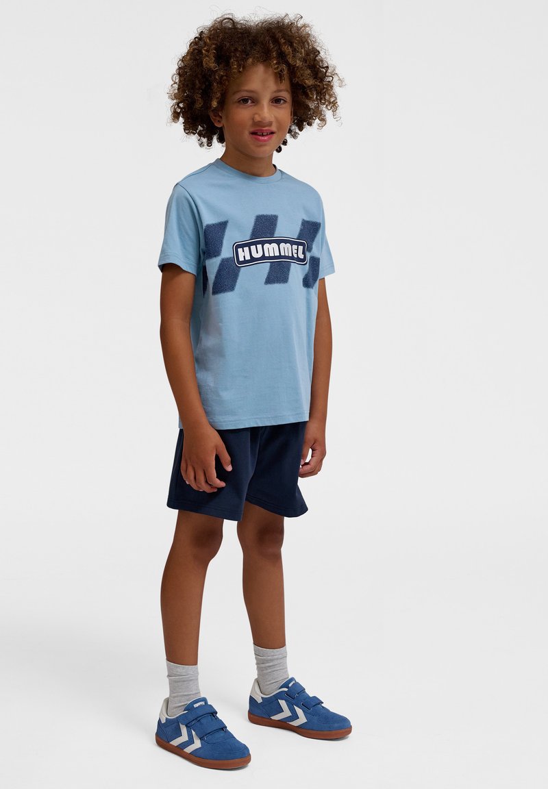 Lyseblå T-shirt med en struktureret mørkeblå logo og mønstrede detaljer, parret med marineblå shorts og blå sko med hvide striber.