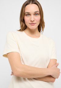 Femme aux cheveux châtain clair et aux yeux bleus, portant un t-shirt crème uni à manches courtes, les bras croisés, sur un fond blanc.